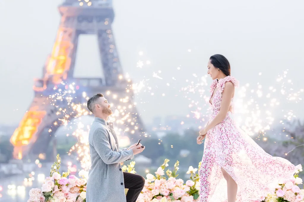 Breathtaking Paris proposal at Trocadero with rare half-lit Eiffel Tower, floral design, fireworks fountains, and gentleman on one knee proposing to woman in flowing pink dress at sunrise.