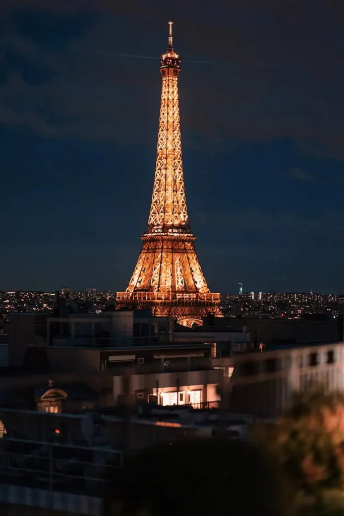 Illuminated Eiffel Tower at blue hour viewed from a private residential rooftop terrace in Paris, with city rooftops in the foreground.