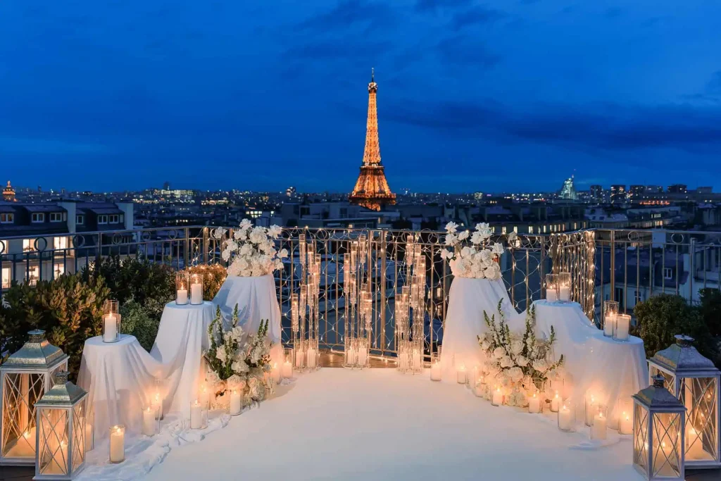 Luxury candlelit Paris proposal setup on residential rooftop terrace at blue hour with illuminated Eiffel Tower, white carpet aisle, romantic candles, glass lanterns, and white rose arrangements by Kiss Me in Paris.