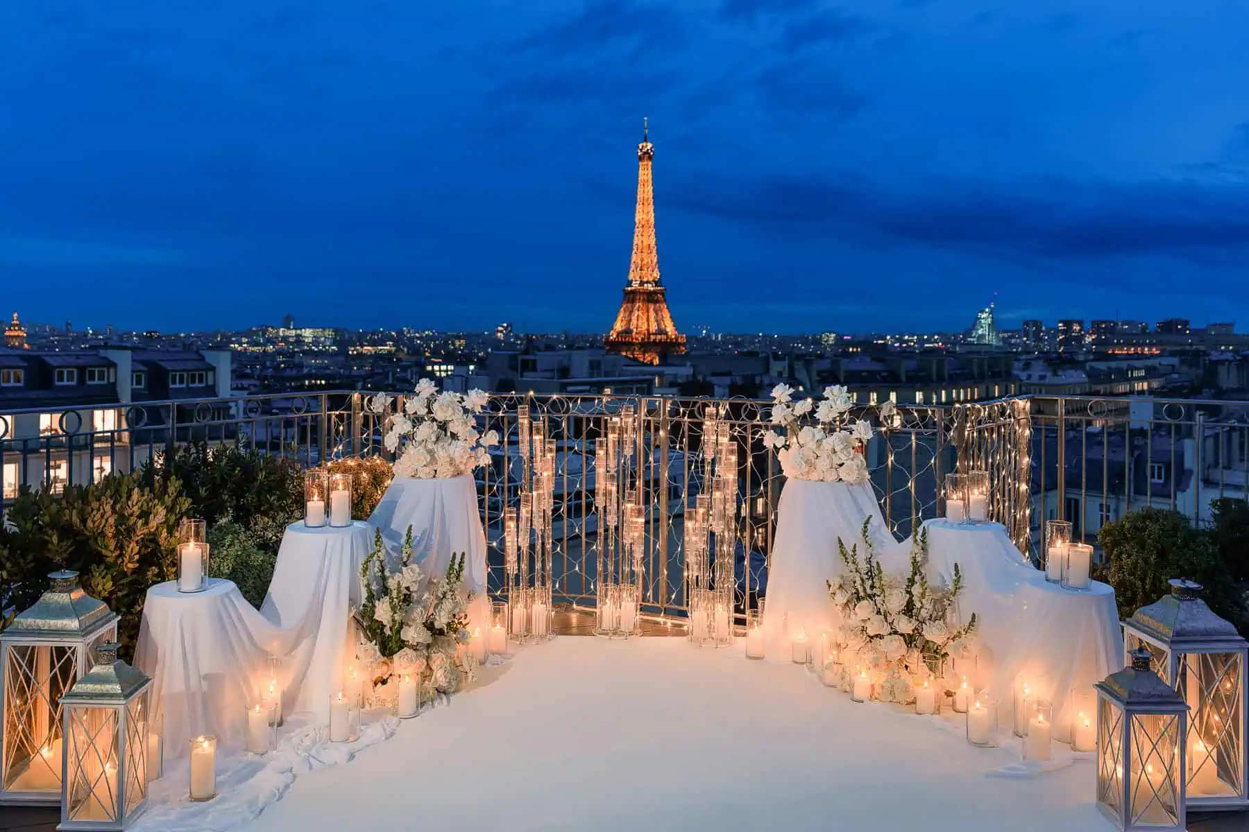 Luxury candlelit Paris proposal setup on residential rooftop terrace at blue hour with illuminated Eiffel Tower, white carpet aisle, romantic candles, glass lanterns, and white rose arrangements by Kiss Me in Paris.