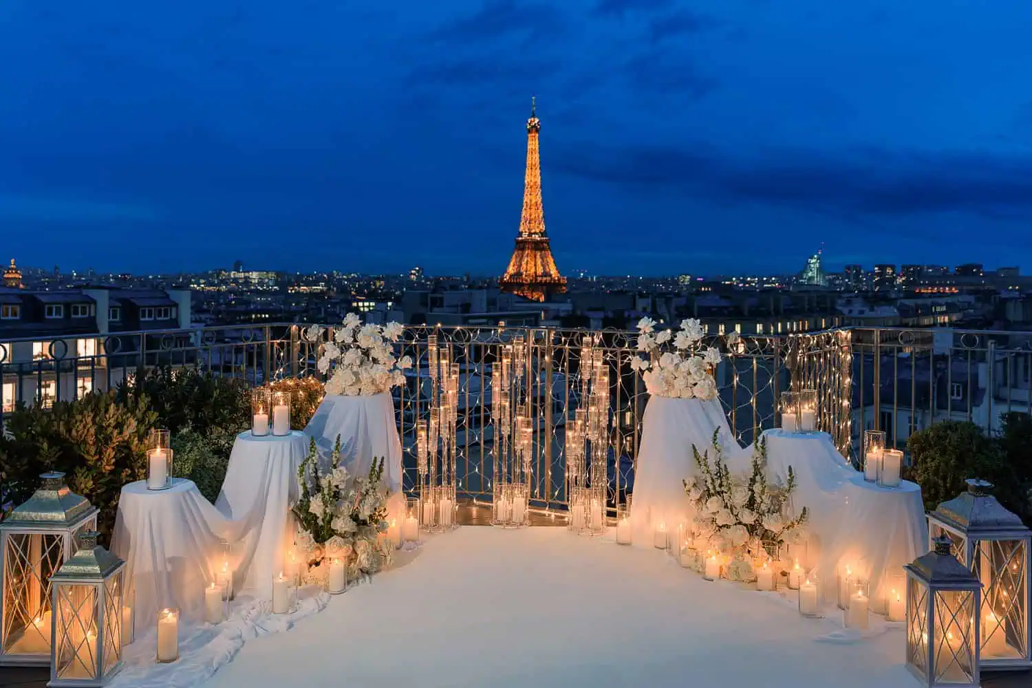 Luxury candlelit Paris proposal setup on residential rooftop terrace at blue hour with illuminated Eiffel Tower, white carpet aisle, romantic candles, glass lanterns, and white rose arrangements by Kiss Me in Paris.