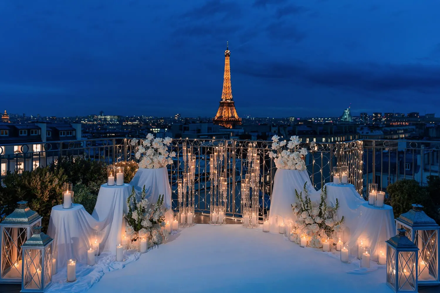 Luxury candlelit proposal setup on Paris rooftop terrace at blue hour with illuminated Eiffel Tower, white carpet aisle, pillar candles, glass lanterns, and white rose arrangements by Kiss Me in Paris.