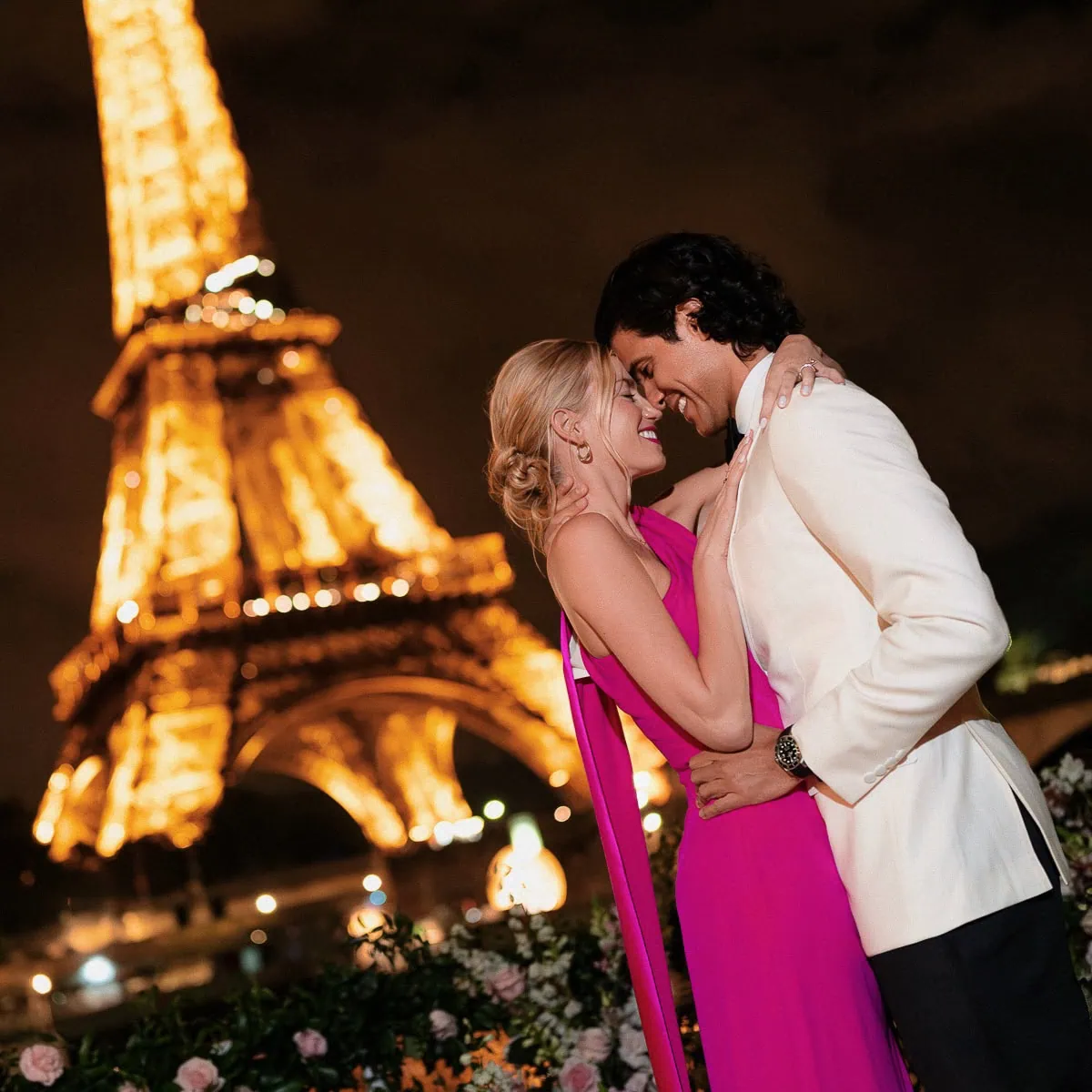 Couple embracing after a Paris proposal with the Eiffel Tower illuminated at night, woman in fuchsia gown, man in white dinner jacket, roses in foreground