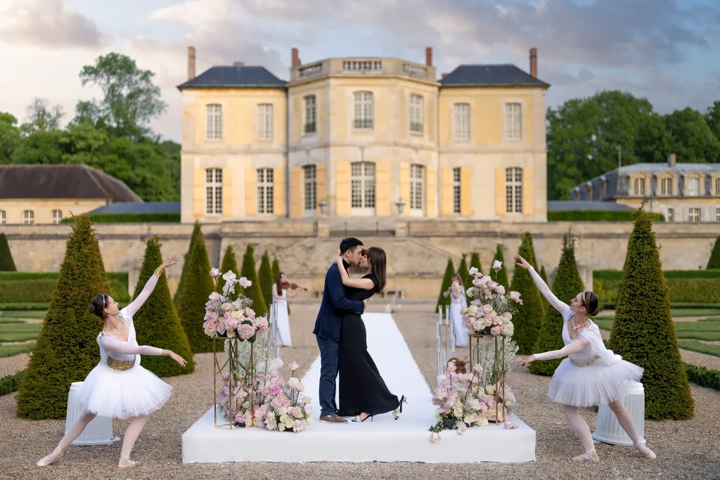 Couple embracing on a white platform in the formal gardens of Château de Villette, flanked by ballet dancers and pink floral arrangements, with the château façade at dusk