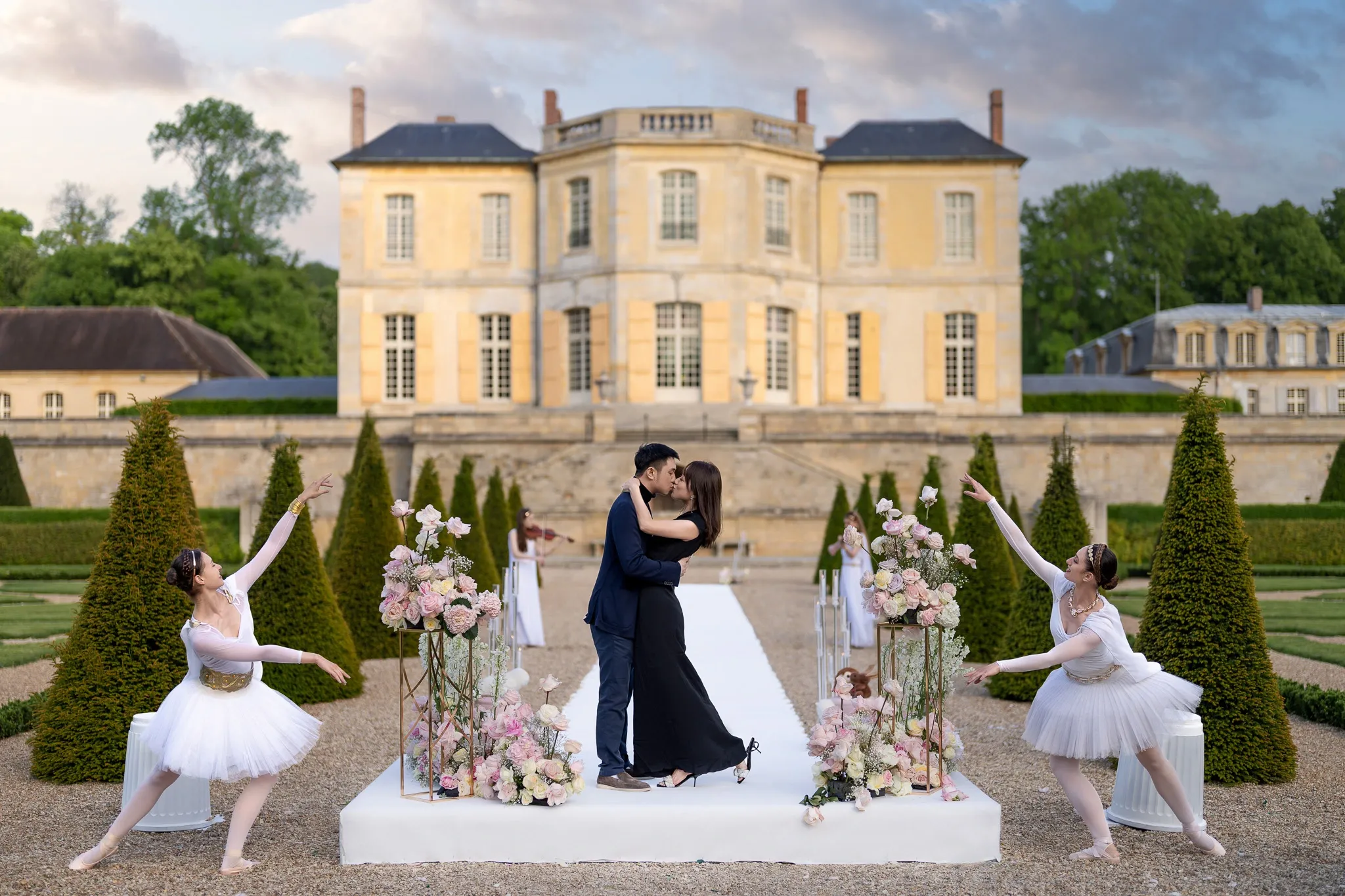 Couple embracing on a white platform in the formal gardens of Château de Villette, flanked by ballet dancers and pink floral arrangements, with the château façade at dusk