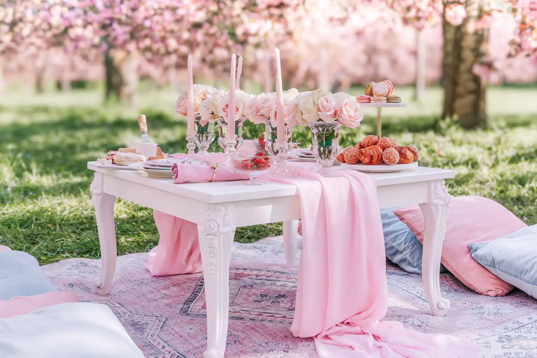 Luxury picnic proposal setup at Parc de Sceaux under cherry blossom trees — white ornate table, pink roses, crystal candelabras, macarons, croissants, and floor cushions on a pink rug
