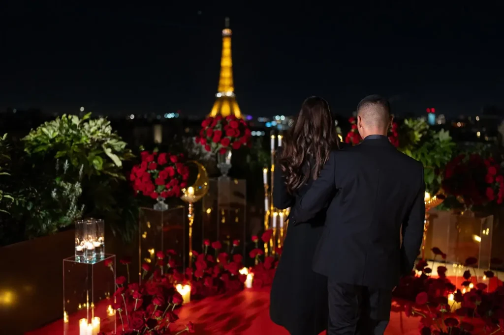 Couple approaching the proposal setup on the Peninsula Paris rooftop at night — red roses, candlelight, crescent moon decor, and the illuminated Eiffel Tower ahead