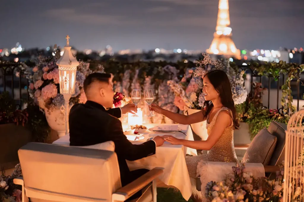Newly engaged couple toasting with champagne at a candlelit rooftop dinner table in Paris — pink hydrangeas and wisteria surrounding them, Eiffel Tower illuminated at night in the background