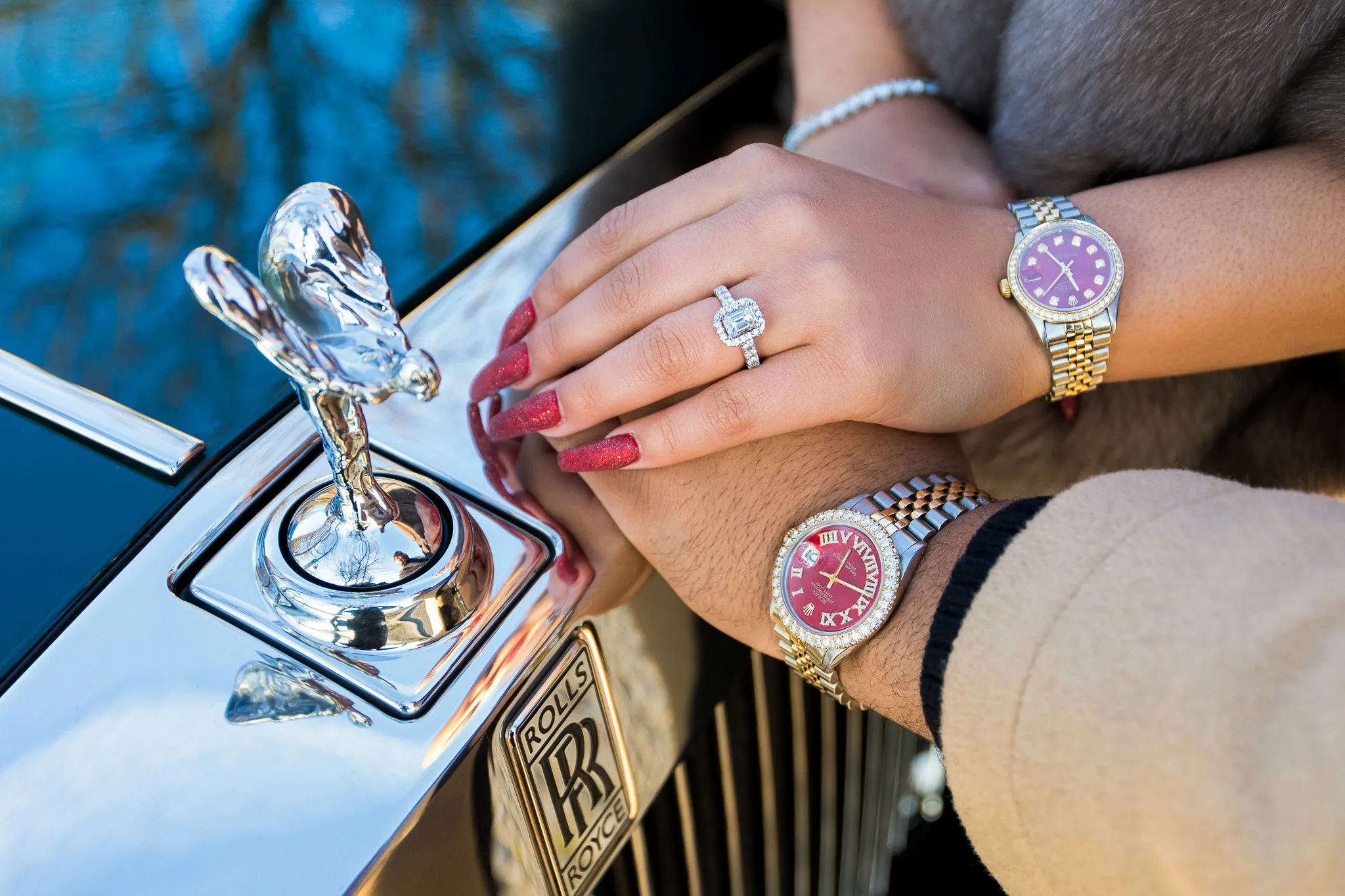 Newly engaged couple's hands resting on a Rolls-Royce hood ornament showing a diamond engagement ring and matching Rolex watches, custom luxury proposal produced by Kiss Me in Paris