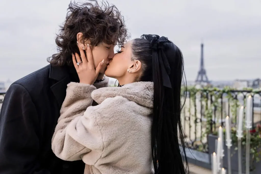 First kiss after a Paris proposal on a rooftop terrace — newly engaged couple kissing with the Eiffel Tower in the background, diamond ring visible on her hand