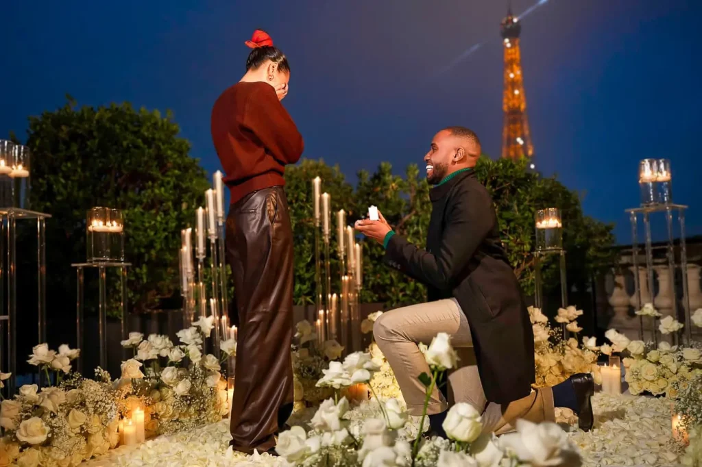 Man on one knee proposing at the Shangri-La Paris terrace at night — open ring box presented, white roses and candles surrounding the couple, Eiffel Tower lit in the background
