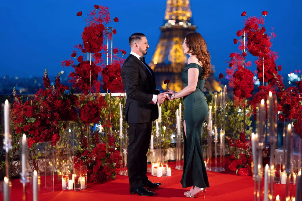 Couple standing in proposal positioning on a Paris rooftop — he on her left, she on his right, hands held facing the Eiffel Tower, surrounded by red roses and candles at night