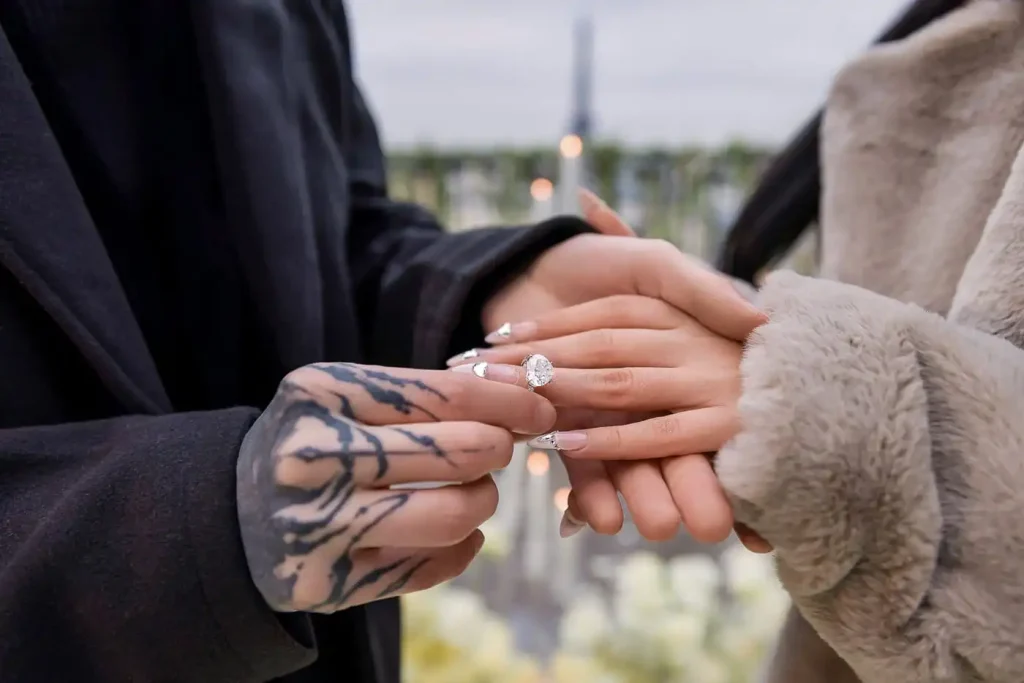 Close-up of the ring exchange during a Paris proposal — tattooed hand sliding a pear-cut diamond engagement ring onto her fourth finger, Eiffel Tower blurred in the background