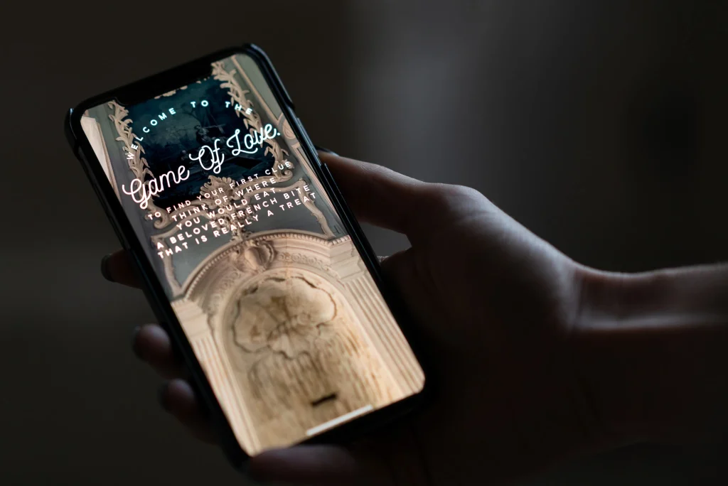 Hand holding a phone displaying a custom "Game of Love" scavenger hunt clue screen, part of a multi-stop proposal experience produced by Kiss Me in Paris at Château de Villette