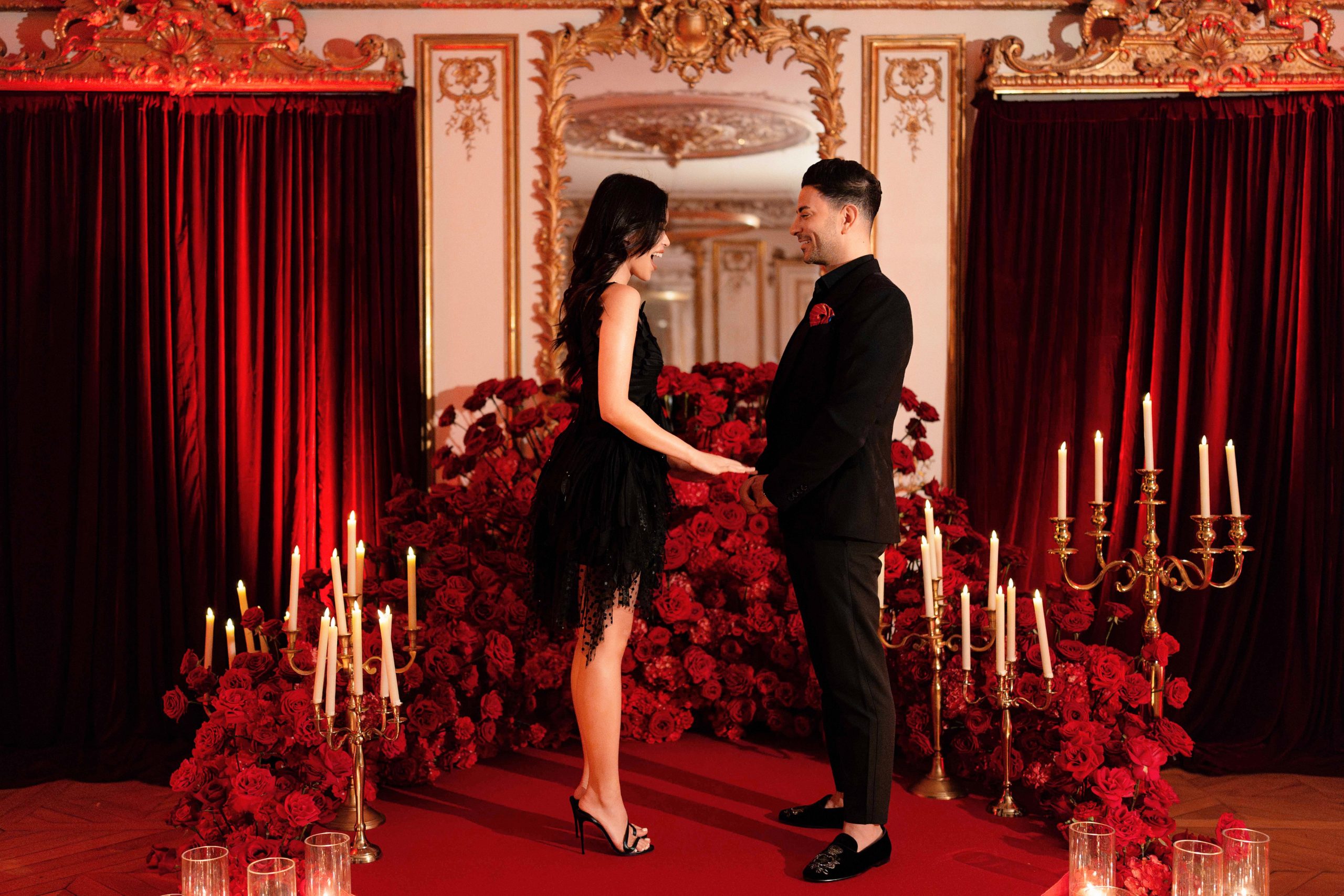 Marriage proposal speech inside a Paris palace salon — the couple standing parallel and facing each other holding hands, she in a black tulle cocktail dress laughing warmly and he in a black suit speaking, surrounded by a curving red-rose floral installation, gold multi-arm candelabras with tall ivory tapers, heavy red velvet drapery, and a gilt rococo mirror above a chevron parquet floor