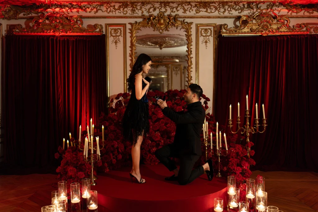 Man proposing on one knee on a red carpet circle surrounded by hundreds of red roses, gold candelabras and candlelight in an ornate Parisian salon, styled venue reveal proposal produced by Kiss Me in Paris