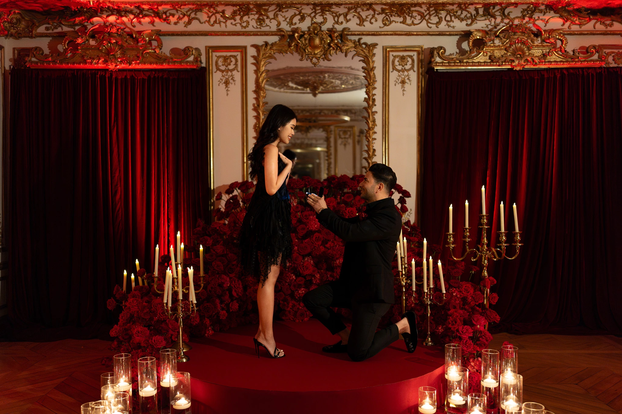 Man proposing on one knee on a red carpet circle surrounded by hundreds of red roses, gold candelabras and candlelight in an ornate Parisian salon, styled venue reveal proposal produced by Kiss Me in Paris