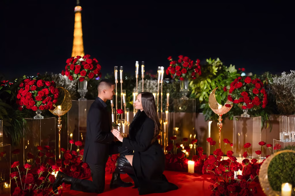 Nighttime marriage proposal at the Peninsula Paris Secret Table rooftop, the Eiffel Tower illuminated in the distance, the gentleman on one knee holding a single red rose and his partner kneeling down to meet him at his level, surrounded by hundreds of red roses, tall taper candles, crescent moon decor, and a red carpet runway