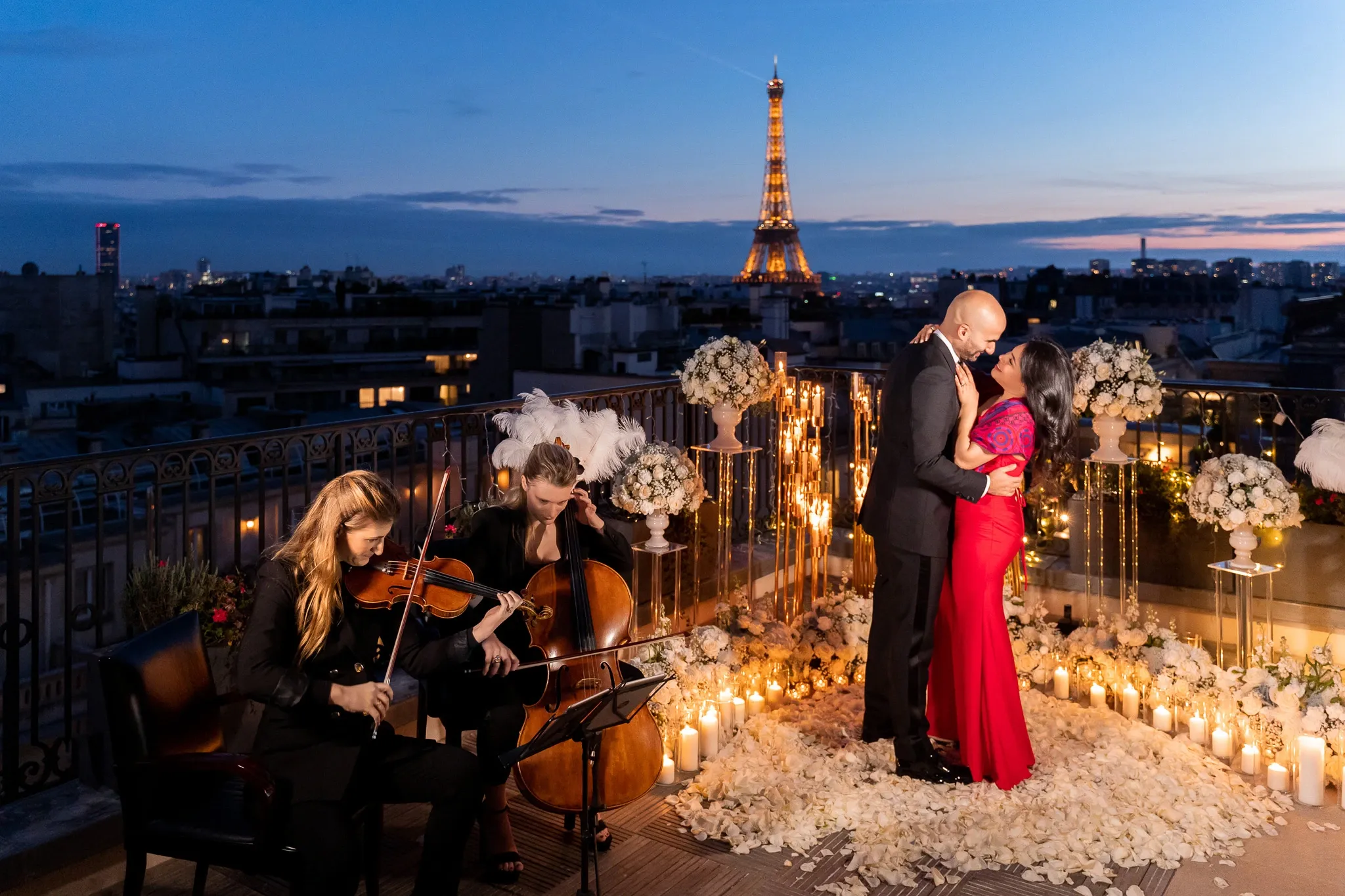 Couple embracing on the Peninsula Paris Secret Table terrace surrounded by white roses, rose petals, candles and gold installations with the illuminated Eiffel Tower at blue hour, proposal produced by Kiss Me in Paris