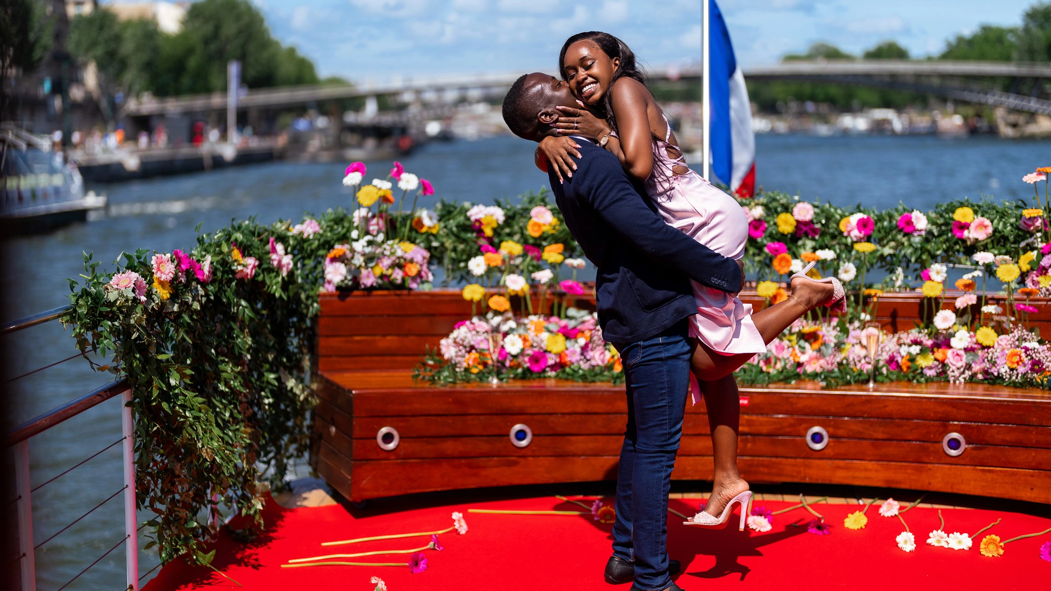 Couple celebrating on a privately chartered yacht on the Seine in Paris — red carpet deck covered in colourful florals, French flag, river and bridge in background