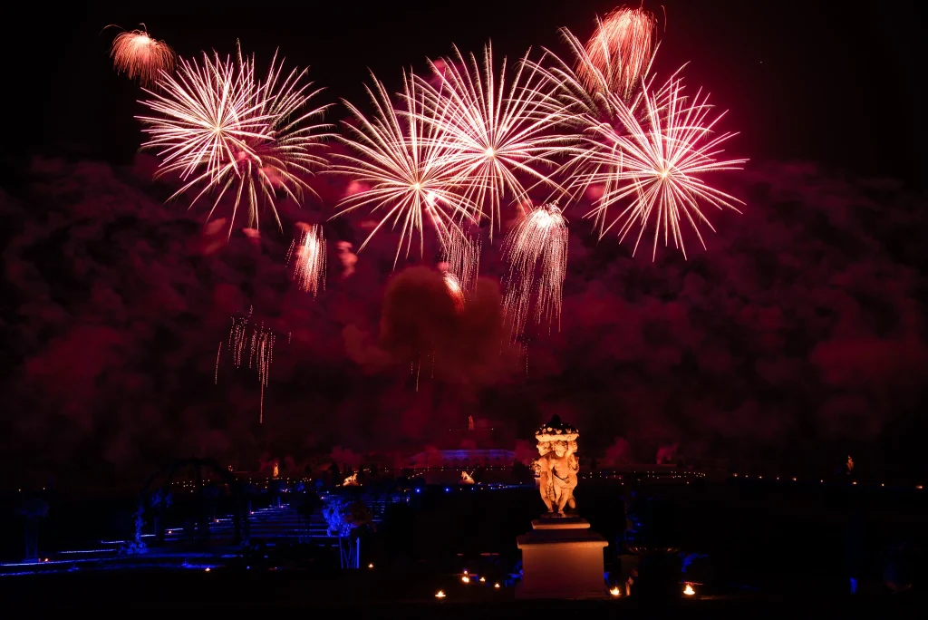 Fireworks display over Vaux-le-Vicomte château gardens at night — classical stone statuary, blue-lit formal gardens, candlelit pathways and crowds below