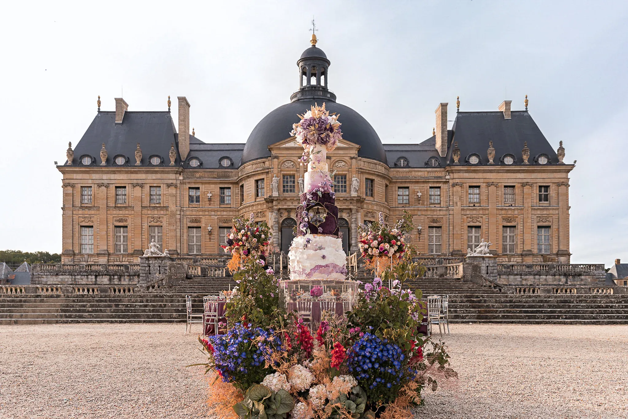 Towering multi-tier purple and white celebration cake surrounded by wildflower arrangements and ghost chairs in front of the Vaux-le-Vicomte château façade, luxury proposal produced by Kiss Me in Paris