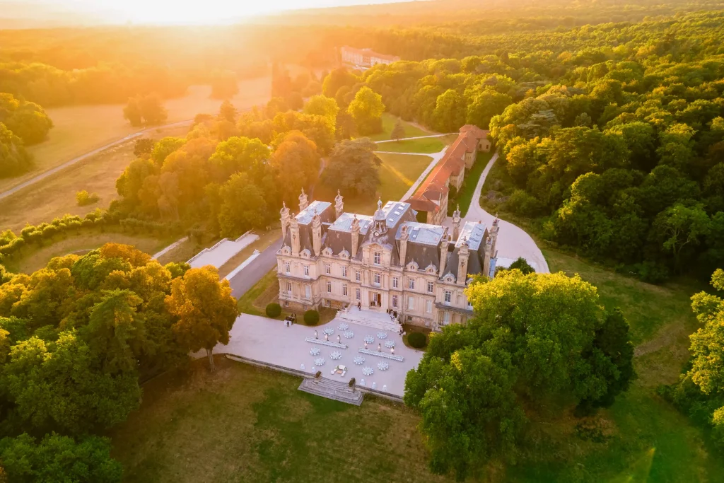 Aerial view of Château Saint-Martin-du-Tertre at golden hour, with 240 wedding guests on the gravel apron and outdoor dinner tables set in front of the lit limestone façade, Val-d'Oise, France.