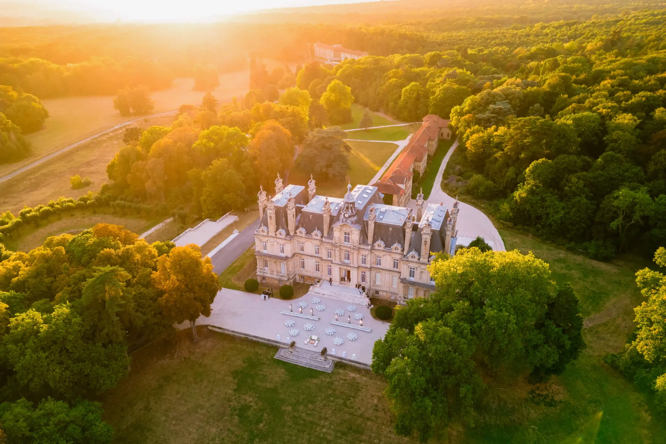 Aerial view of Château Saint-Martin-du-Tertre at golden hour, with 240 wedding guests on the gravel apron and outdoor dinner tables set in front of the lit limestone façade, Val-d'Oise, France.