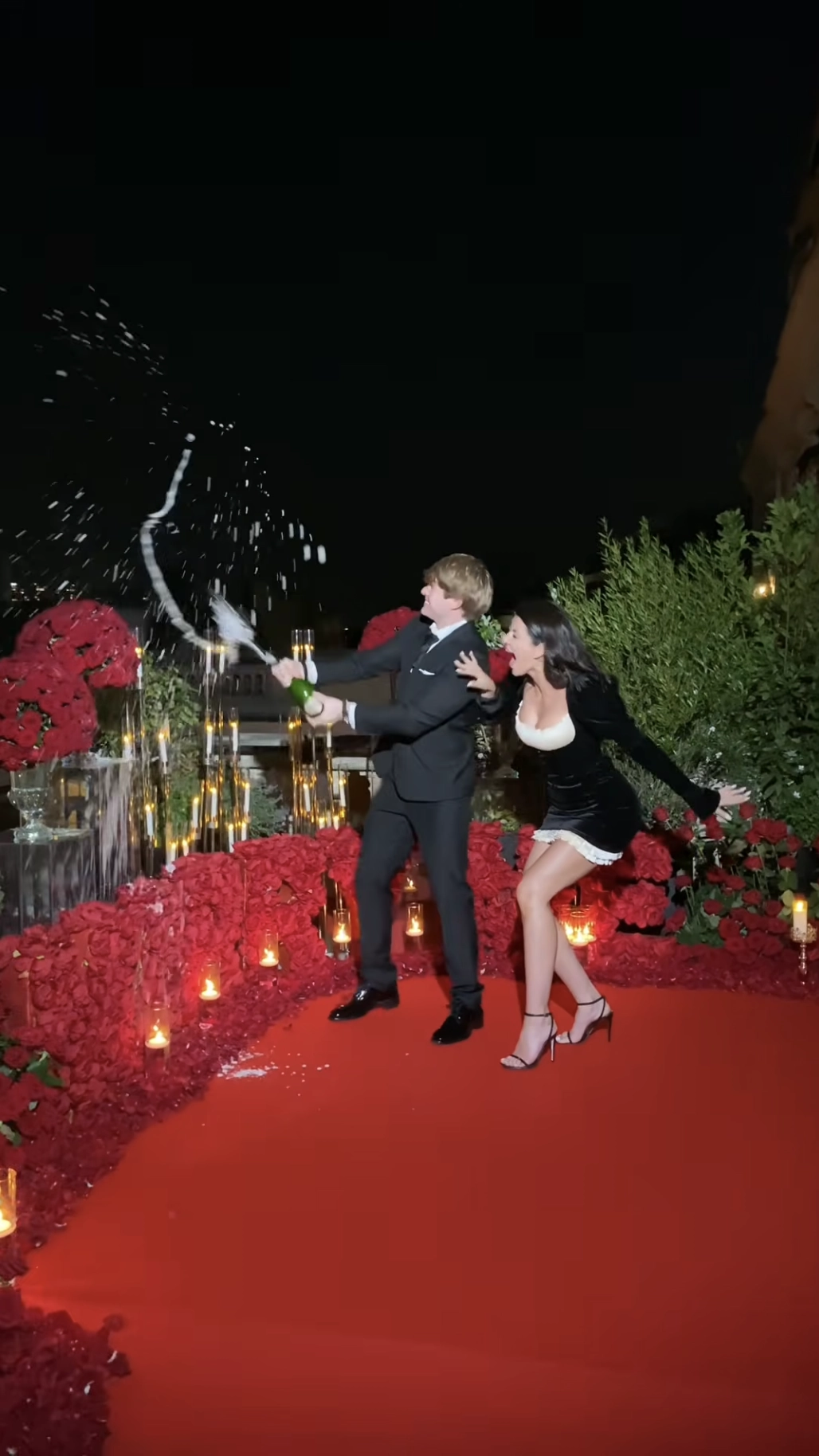 A newly engaged couple popping champagne on a red carpet rooftop terrace at Shangri-La Paris, surrounded by red rose arrangements and string lights at night