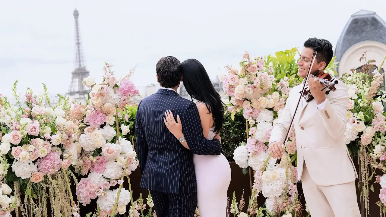 Couple embracing with a view of the Eiffel Tower as a violinist performs beside pink and white florals at The Peninsula Paris