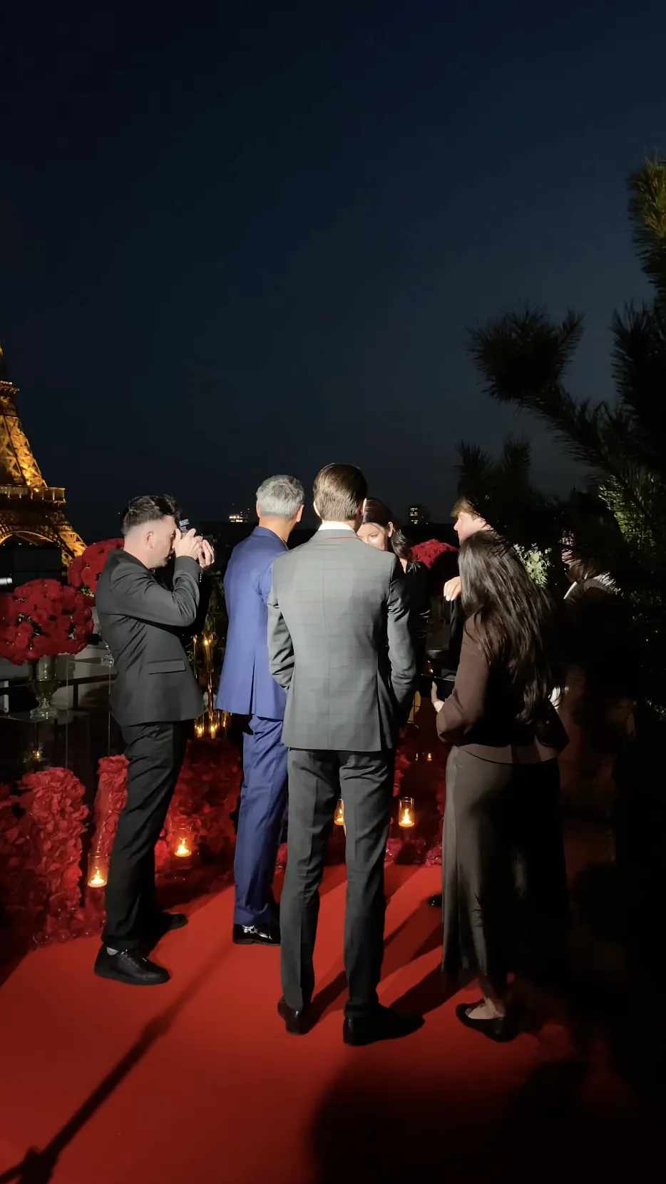Family members gathered on a rooftop terrace at Shangri-La Paris during a proposal, with the illuminated Eiffel Tower in the background and red rose arrangements lining a red carpet