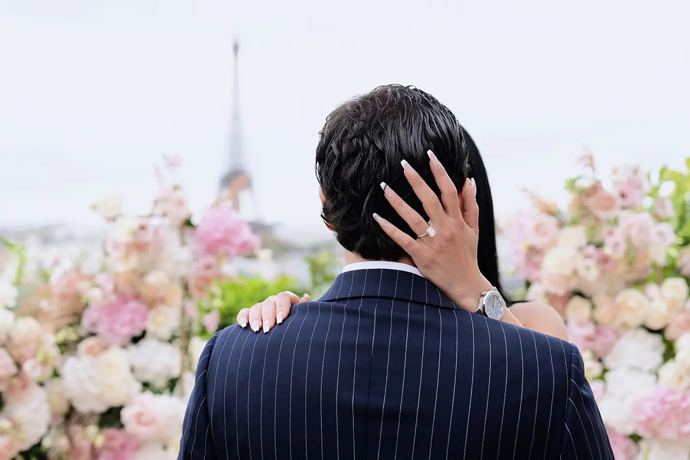 Close-up of an engagement ring on a woman's hand resting on her partner's neck with the Eiffel Tower in the background