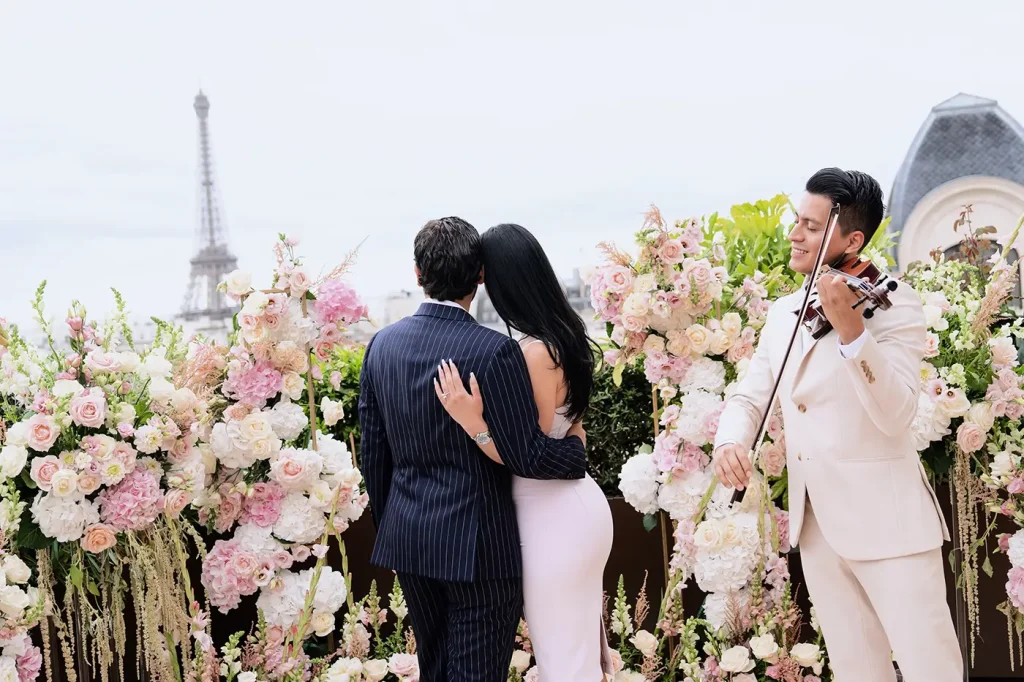 Couple embracing on The Peninsula Paris rooftop as a live violinist performs beside the floral arch