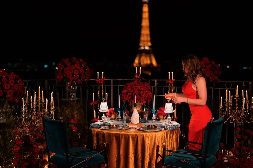 A proposal planner in a red dress standing at a fully set rooftop dinner table in Paris at night, with the illuminated Eiffel Tower visible behind her, surrounded by red roses, gold candelabras, and teal velvet chairs