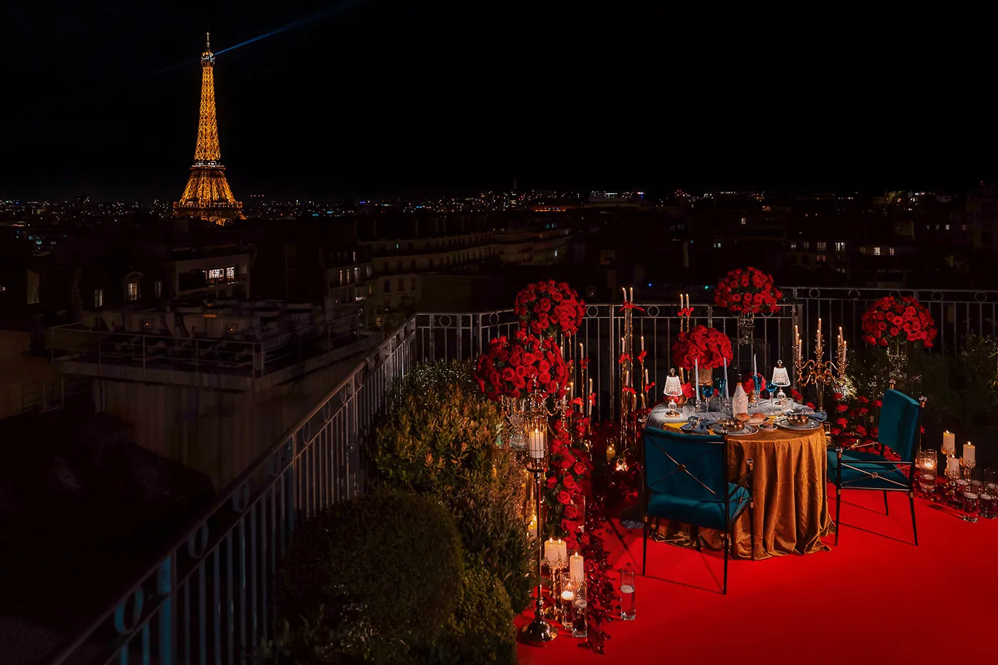 Rooftop dinner table set for a marriage proposal in Paris at night, with the illuminated Eiffel Tower in the background, red roses, gold tablecloth, teal velvet chairs, and candlelight