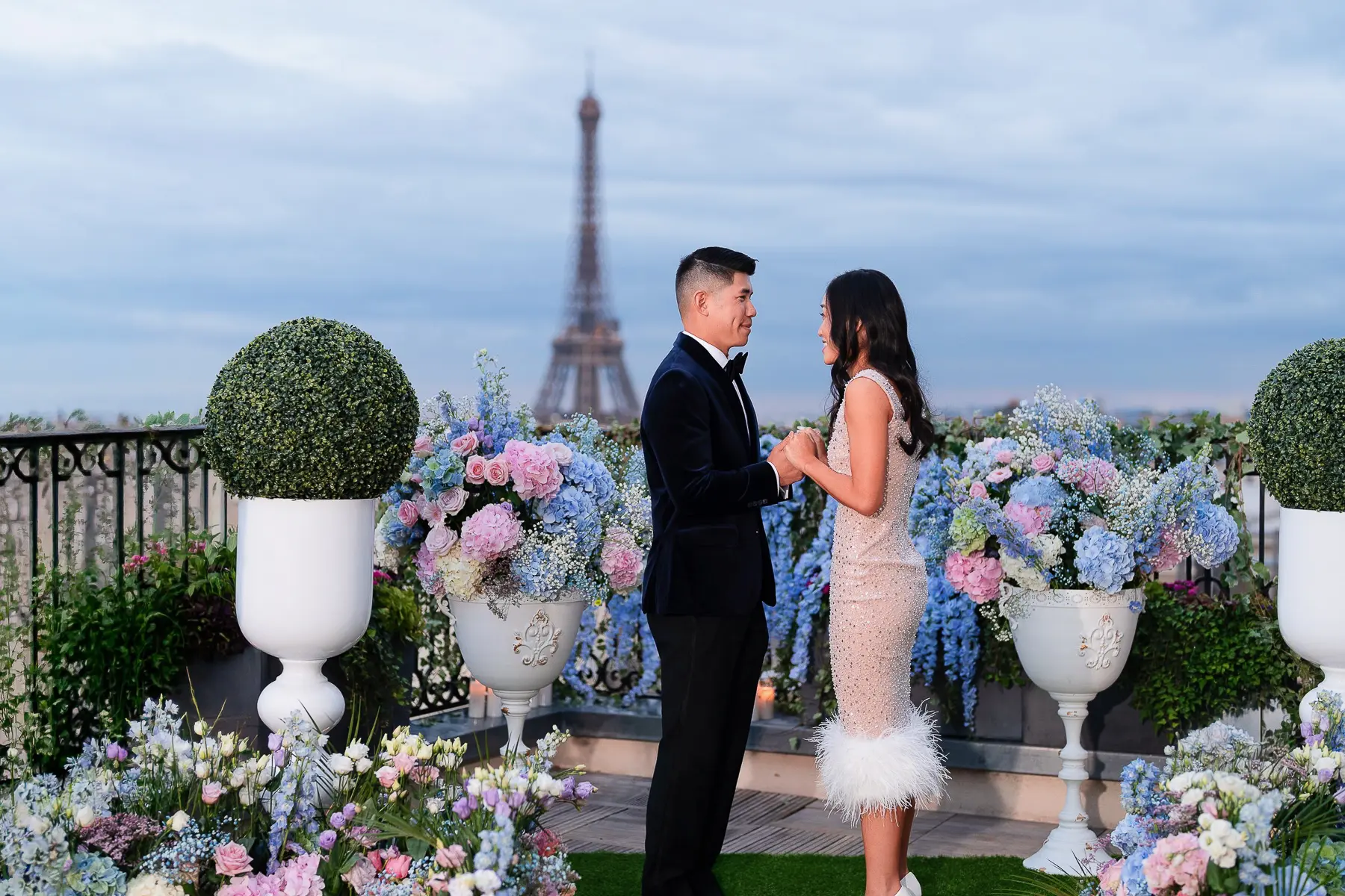 Couple holding hands after a rooftop proposal surrounded by blue, pink, and white floral arrangements with the Eiffel Tower in the background.