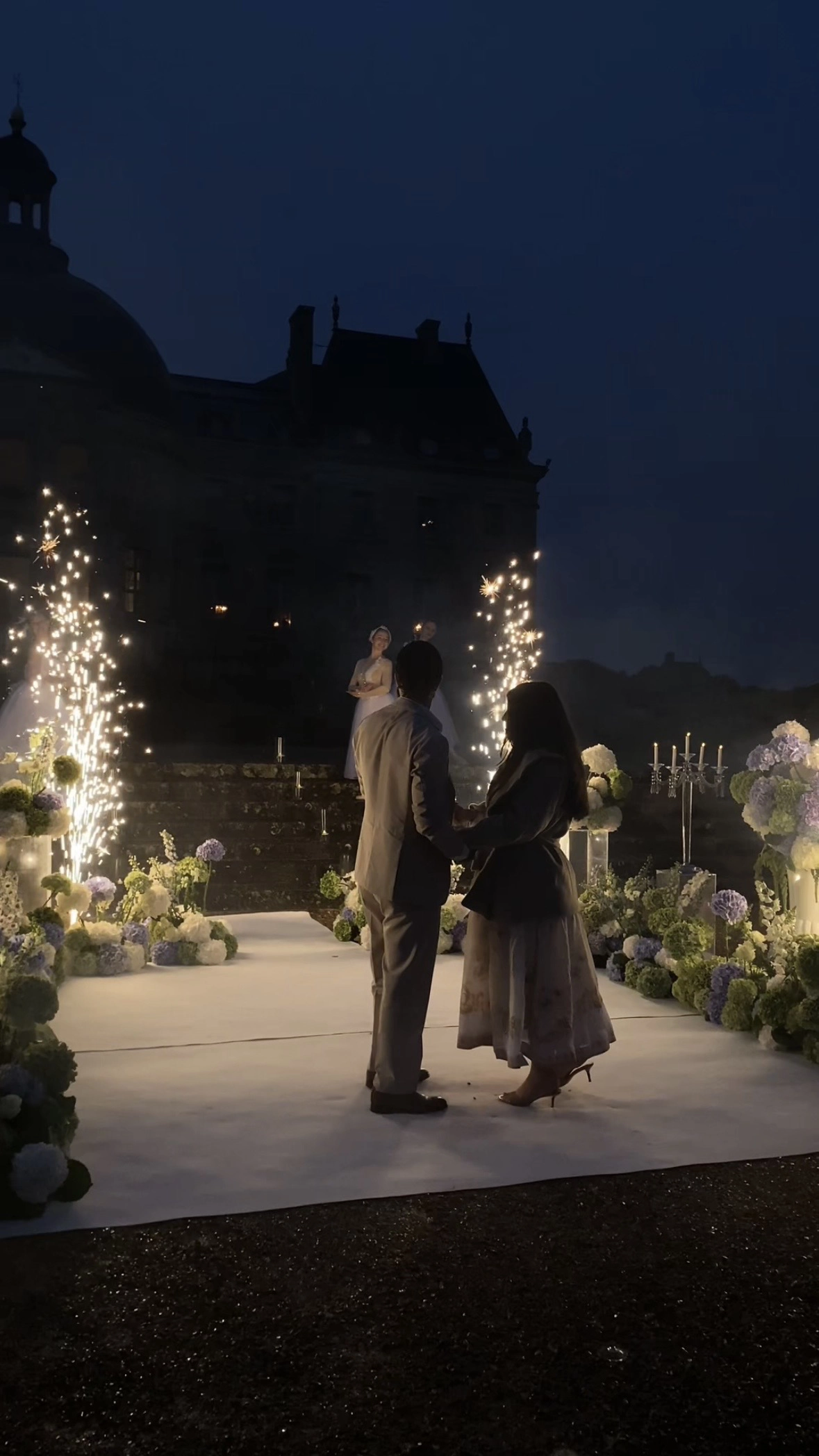 Couple stands together during proposal as sparkler fountains and ballerinas light the château steps behind them