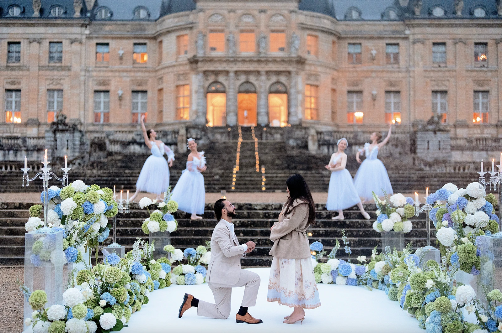 Man on one knee proposing on château steps as ballerinas dance behind him
