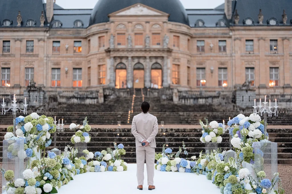 Man holds ring box behind his back while waiting on château steps in the rain