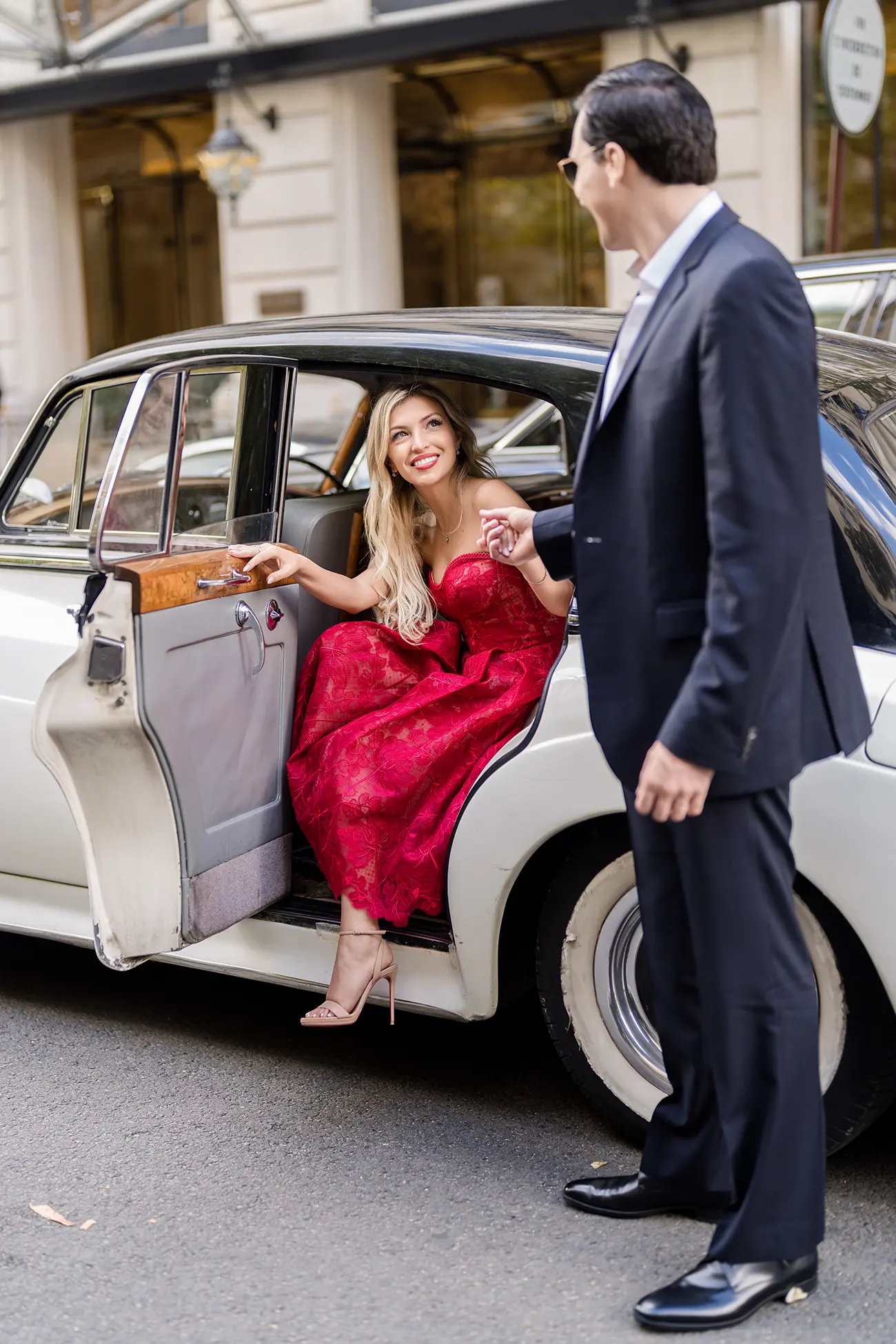 Woman in red dress stepping out of a vintage white car, helped by her partner outside The Peninsula Paris