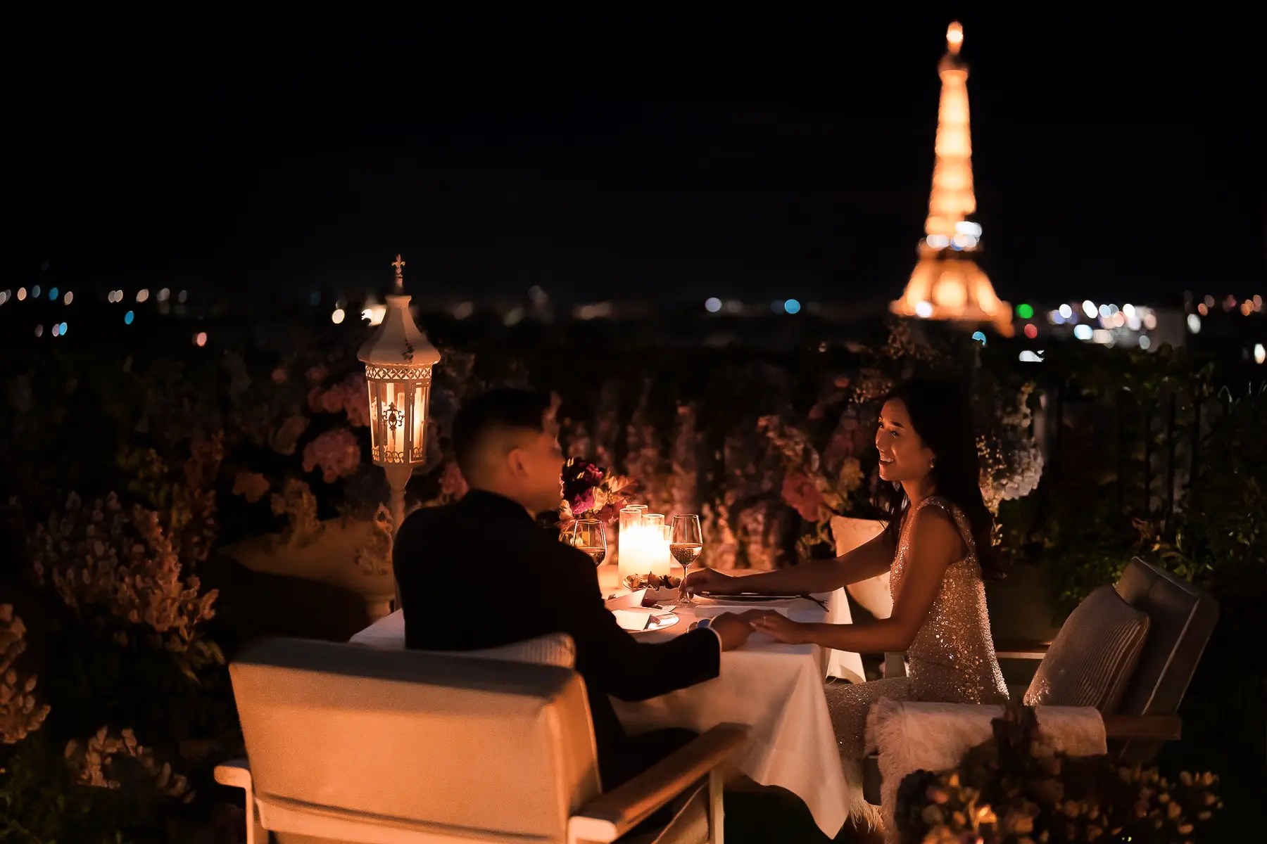 Couple holding hands across a candlelit dinner table on The Peninsula Paris rooftop at night with Eiffel Tower view