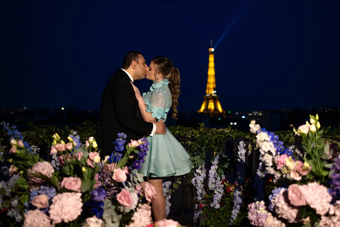 Couple kissing on The Peninsula Paris rooftop at night, floral-covered railing and Eiffel Tower behind them