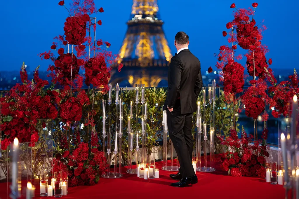 Man in tuxedo standing alone on The Shangri-La Paris terrace surrounded by red roses and candlelight, Eiffel Tower at blue hour behind him