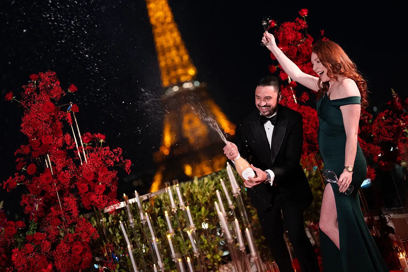 Couple spraying champagne on The Shangri-La Paris terrace, red roses and Eiffel Tower behind them at night