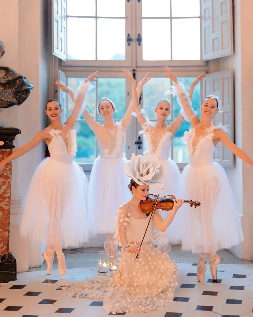 Solo violinist in floral headpiece performs with four ballerinas in front of tall château windows