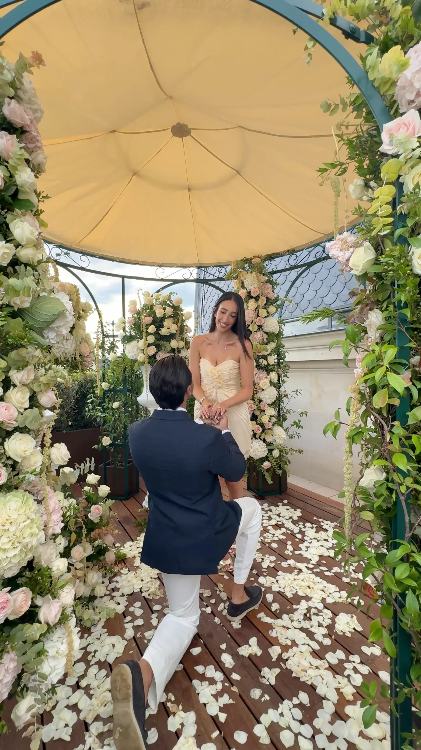 Man kneeling to propose inside a flower-covered gazebo on The Peninsula Paris rooftop, with a woman in a cream dress smiling down at him surrounded by white rose petals scattered across the deck