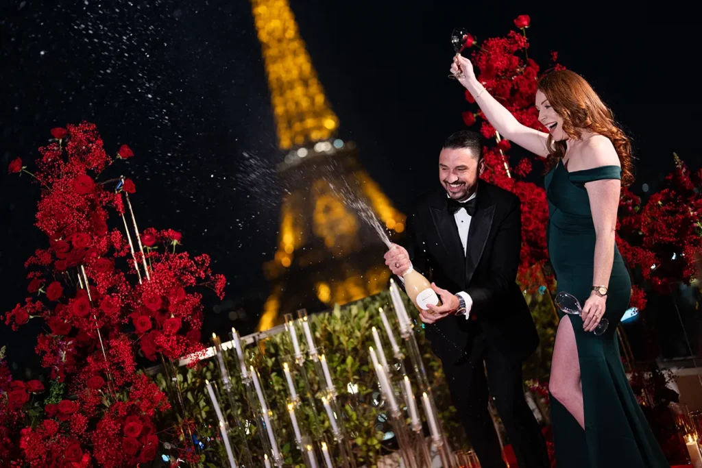 Man popping champagne as his partner raises a glass in celebration on a terrace decorated with red roses and candles, Eiffel Tower lit up behind them