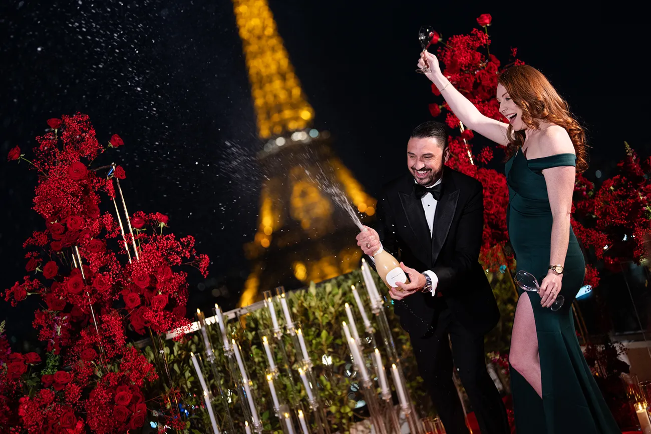 Man popping champagne as his partner raises a glass in celebration on a terrace decorated with red roses and candles, Eiffel Tower lit up behind them