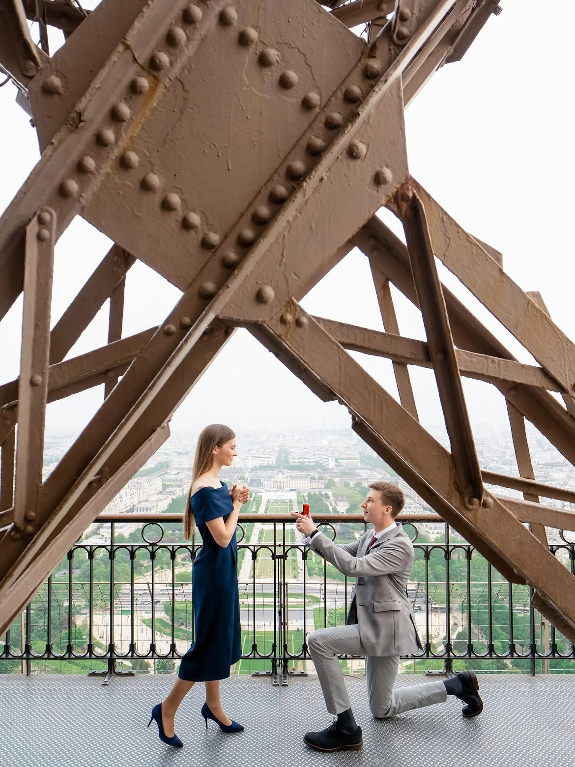 Man kneeling to propose on the Eiffel Tower's second-floor platform, framed by iron beams with Champ de Mars below