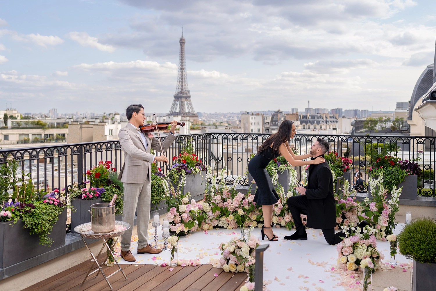 Man kneeling to propose on The Peninsula Paris's Secret Table terrace, with a floral aisle, champagne, and the Eiffel Tower visible over the Paris rooftops